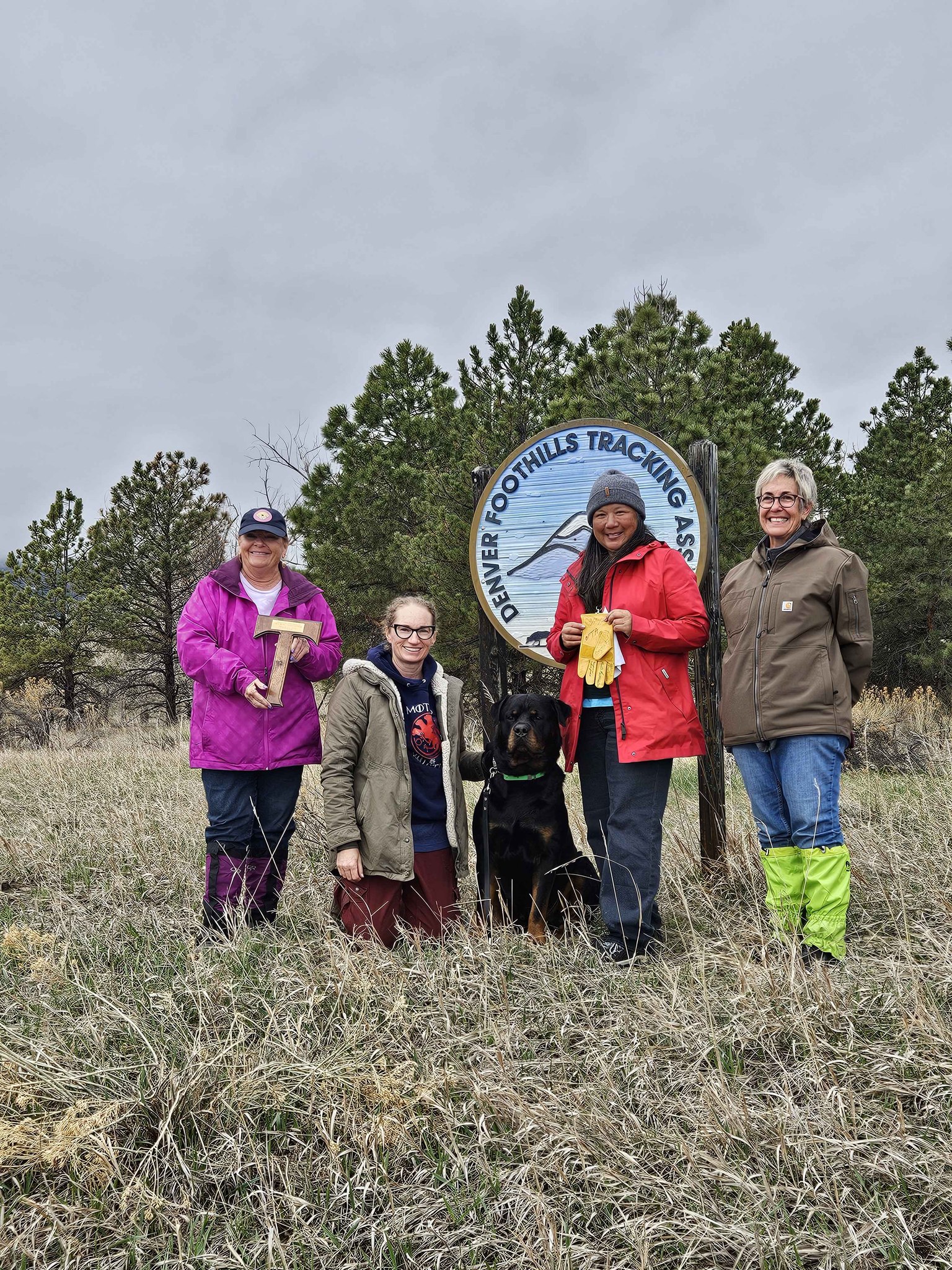 Jill and friends posing alongside her rottweiler, Reve, in front of a sign for the Denver Foothills Tracking Asssociation